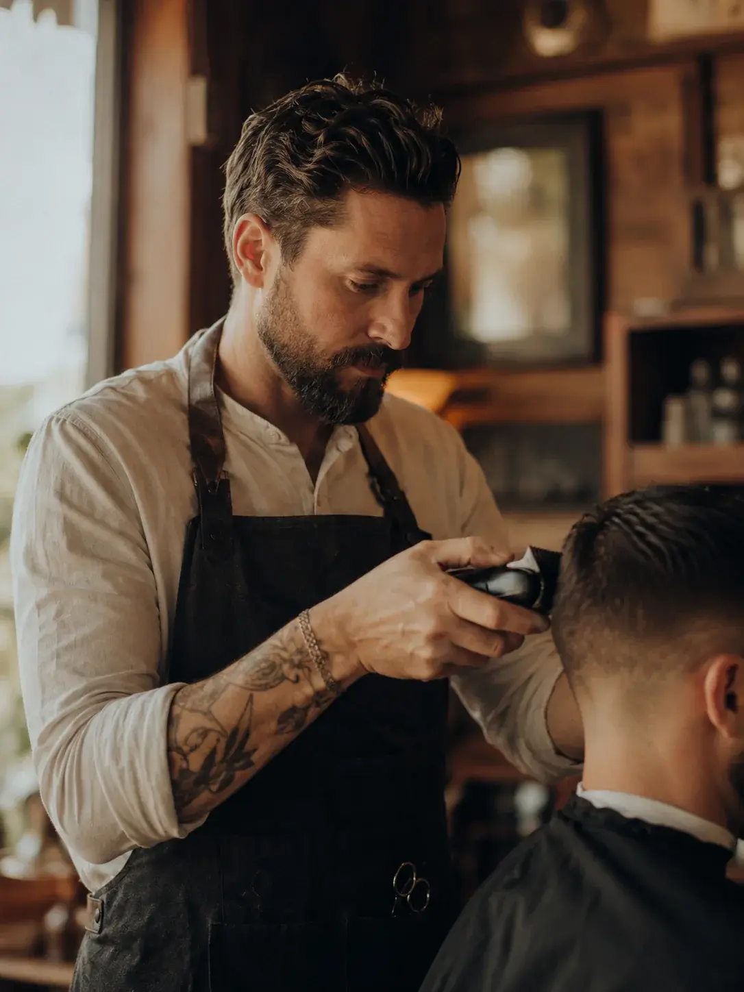 A barber in an apron finishing a client's fade with clippers in a warm, wood-paneled shop